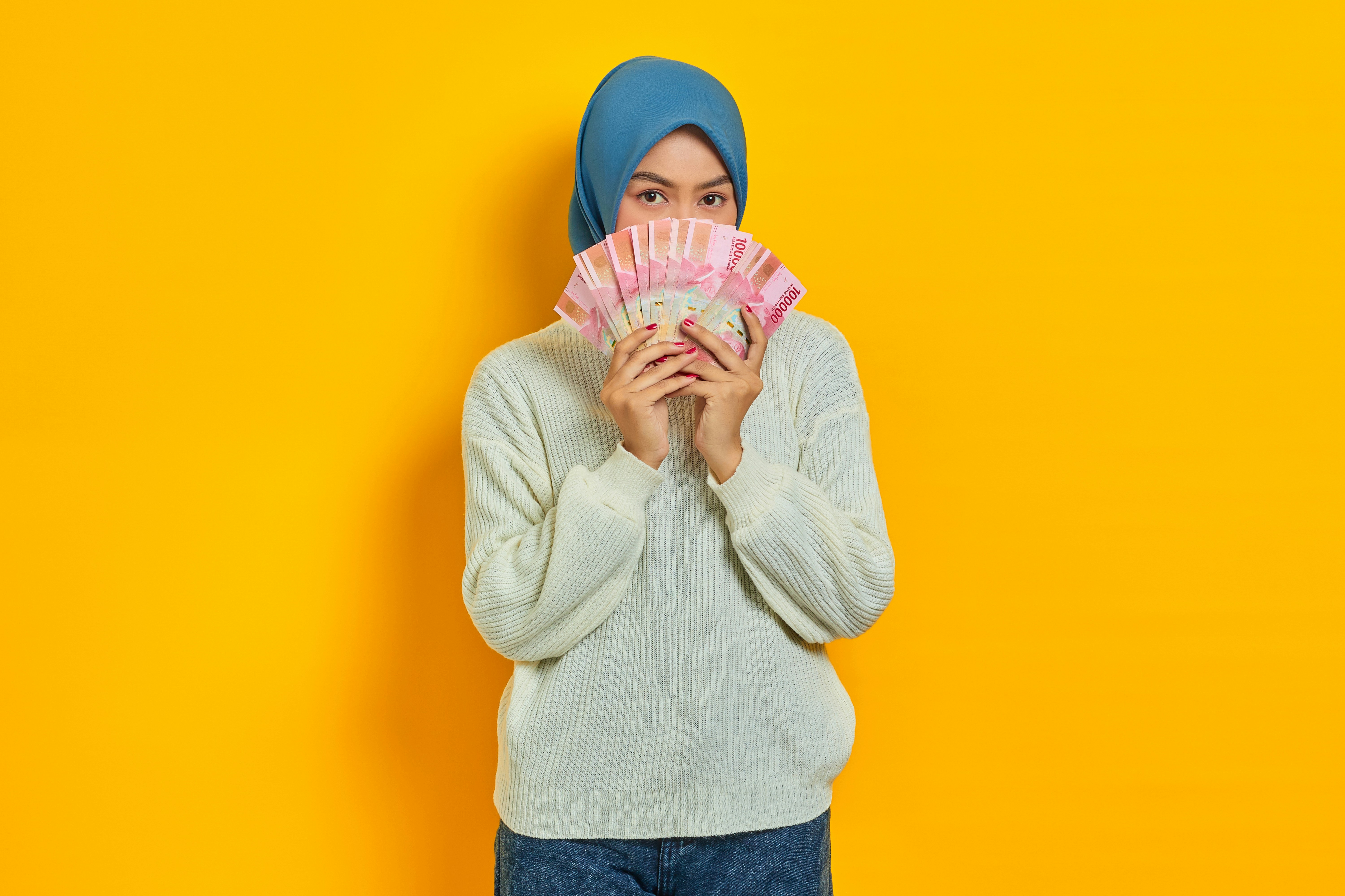 Beautiful Asian Muslim woman in white sweater covering face with cash money in Indonesian rupiah banknotes isolated over yellow background. People religious lifestyle concept